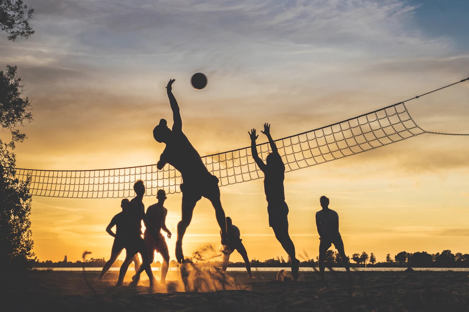 Silhouetted players at a net on sand, a spike and a block against the sunset.